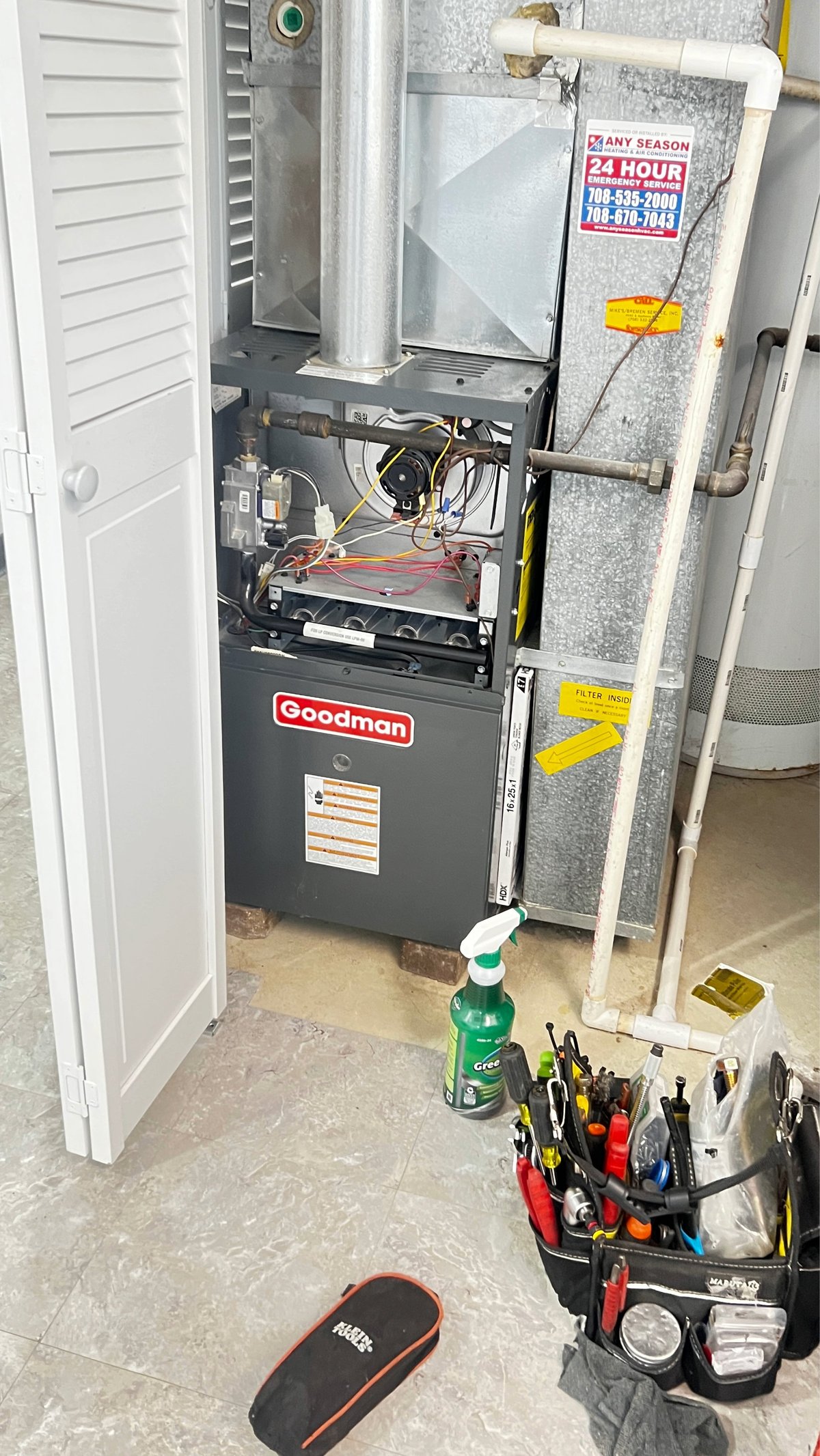 Goodman furnace unit with open panel showing internal components, tools, and equipment in basement utility closet